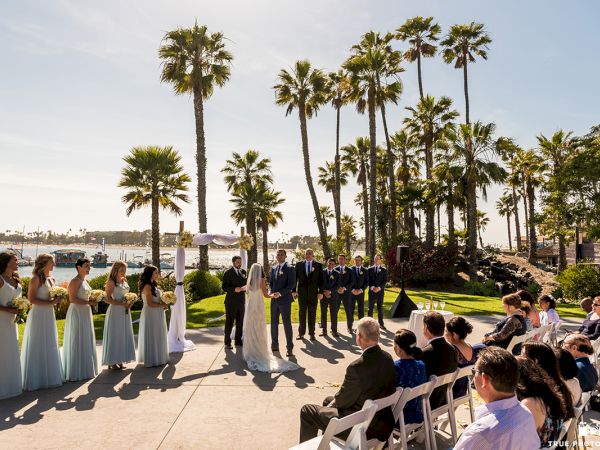 An outdoor wedding ceremony by a waterfront with tall palm trees, bridesmaids in white, groomsmen in suits, and seated guests.