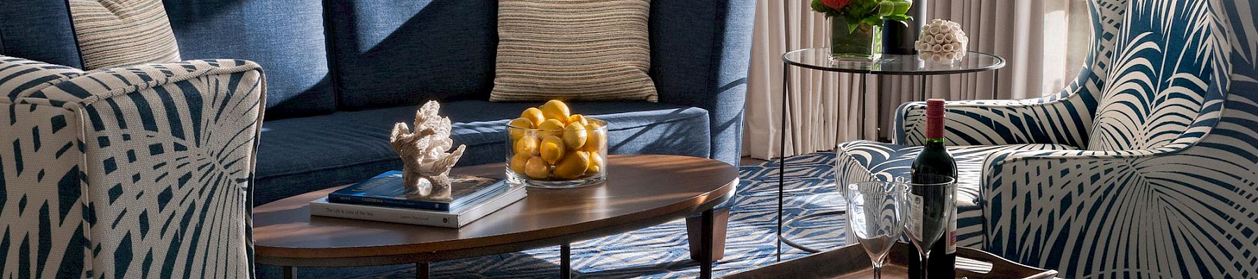 A bright lounge with blue sectional, patterned chairs, a coffee table with grapes, and a small flower-and-wine serving setup.