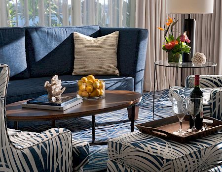 A bright lounge with blue sectional, patterned chairs, a coffee table with grapes, and a small flower-and-wine serving setup.