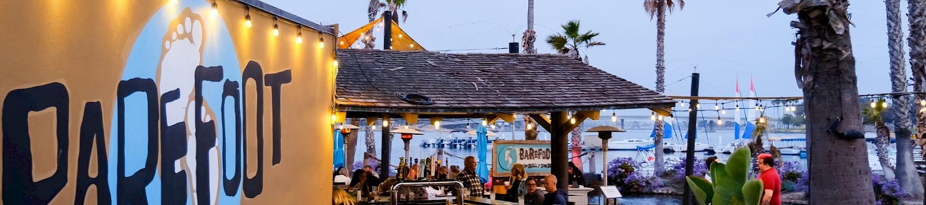Outdoor bar & grill patio with string lights, palm trees, and people by a marina at dusk.