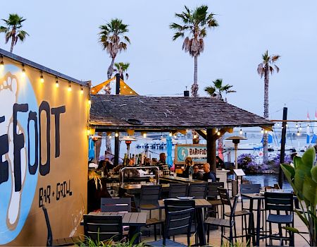 Outdoor bar & grill patio with string lights, palm trees, and people by a marina at dusk.