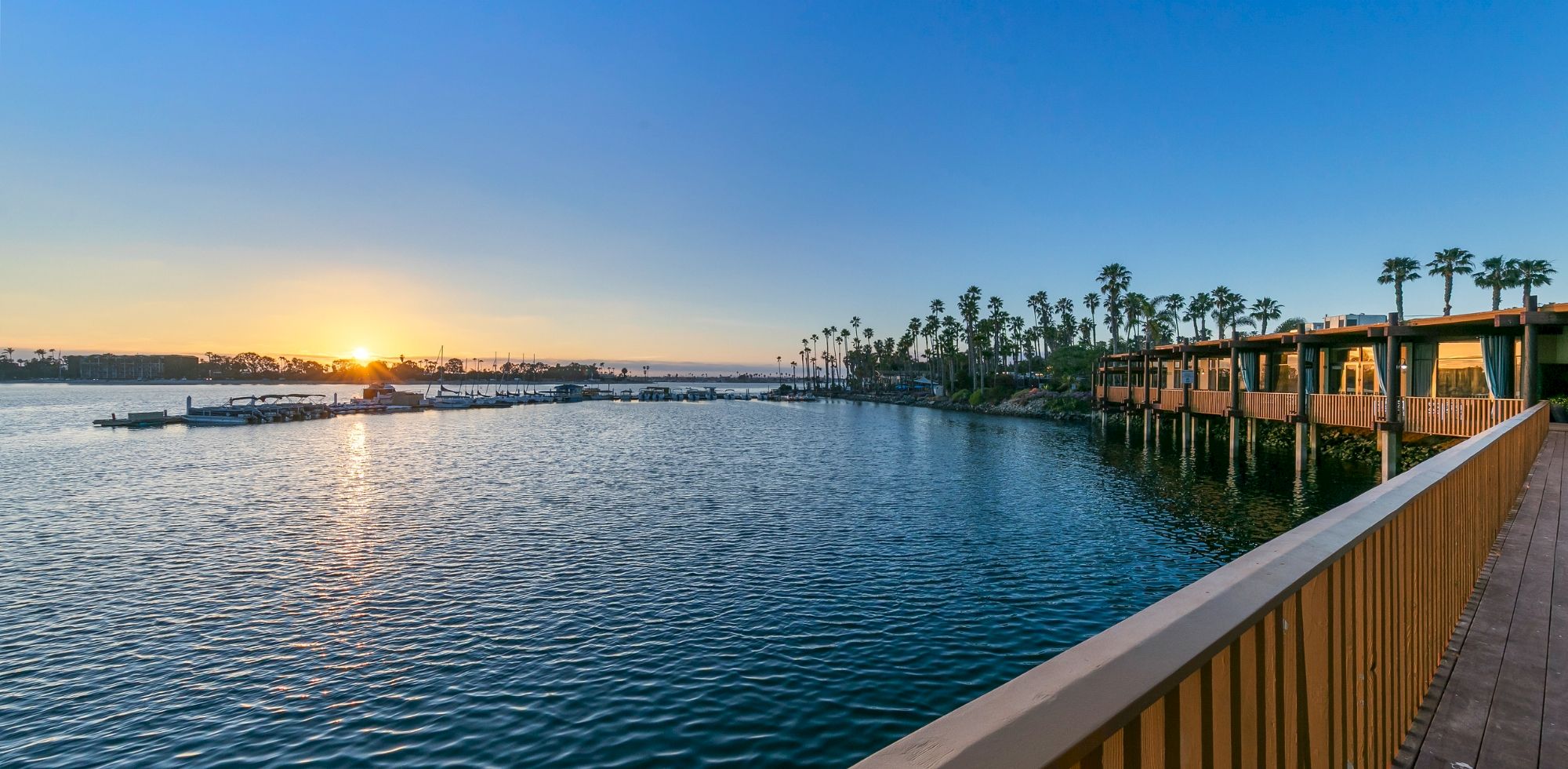Sunrise over a calm harbor with boats, palm trees, and a wooden boardwalk by waterfront buildings, bathing the scene in golden light.