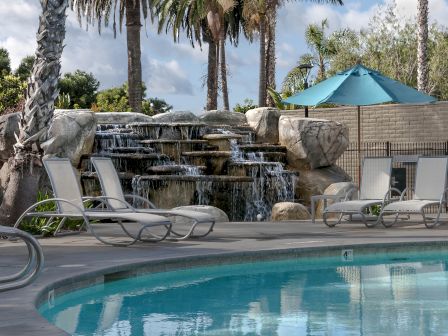 A tropical poolside oasis with cascading waterfalls, palm trees, lounge chairs, and a teal umbrella beside a fenced water feature.