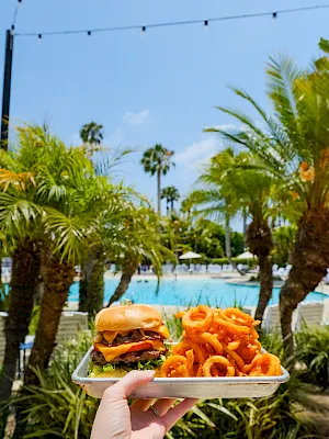 A hand holds a plate with a burger and fries in a sunny poolside garden resort, with palm trees and a blue pool behind.