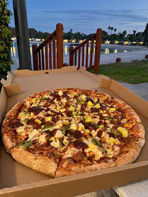 A pepperoni-and-veggie pizza with jalapeño and cheese sits in a cardboard box on a lakeside walkway at dusk.