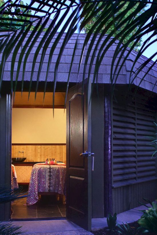 A tropical garden doorway opens to a cozy dining spot with a covered table, framed by lush palms and arching walls.