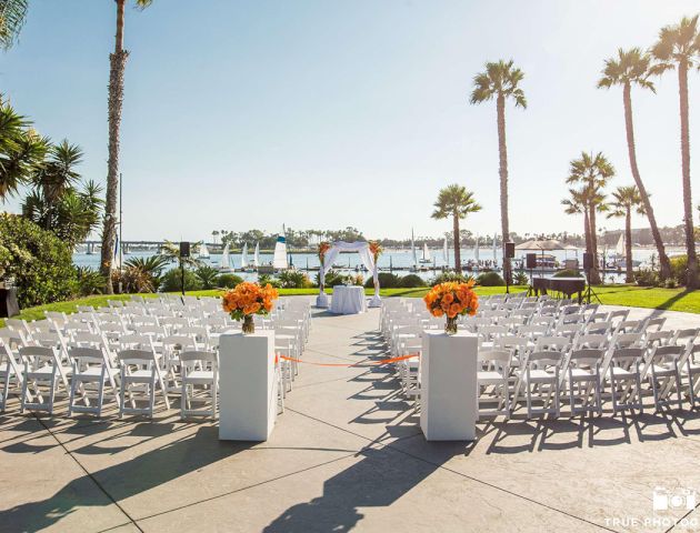 Outdoor wedding ceremony setup with white chairs, orange flower centerpieces, palm trees, and a view of the ocean.