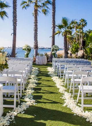 A beach wedding aisle with white chairs, palm trees, and lush white floral bouquets leading to the ocean.