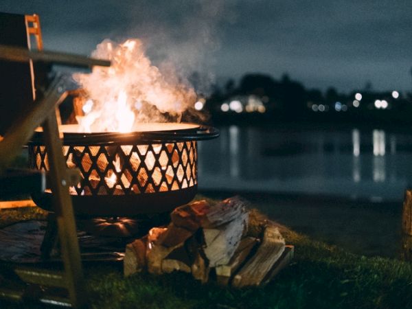 A bright campfire burns in a metal fire pit at night beside scattered logs near dark water and city lights.