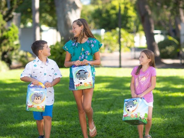 Three kids are smiling in a sunny park while carrying colorful lunchbox bags.