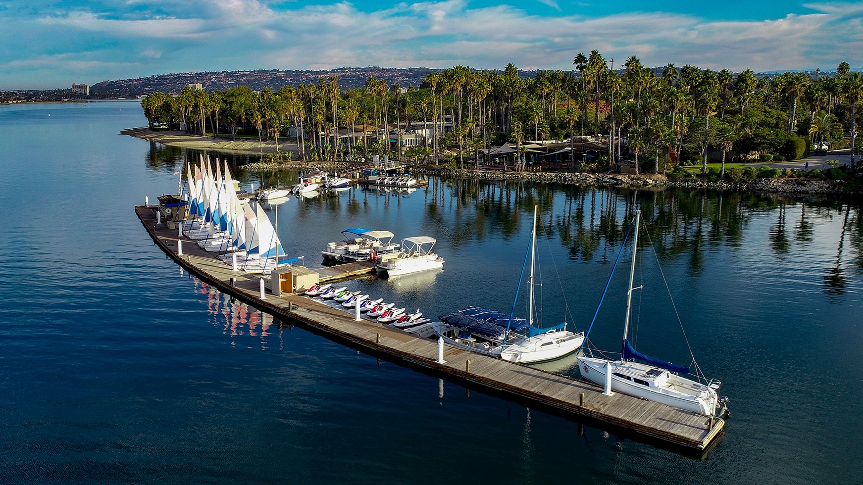 A docked sailboat and yachts sit on a pier beside calm water and a palm-lined waterfront resort under a blue sky.