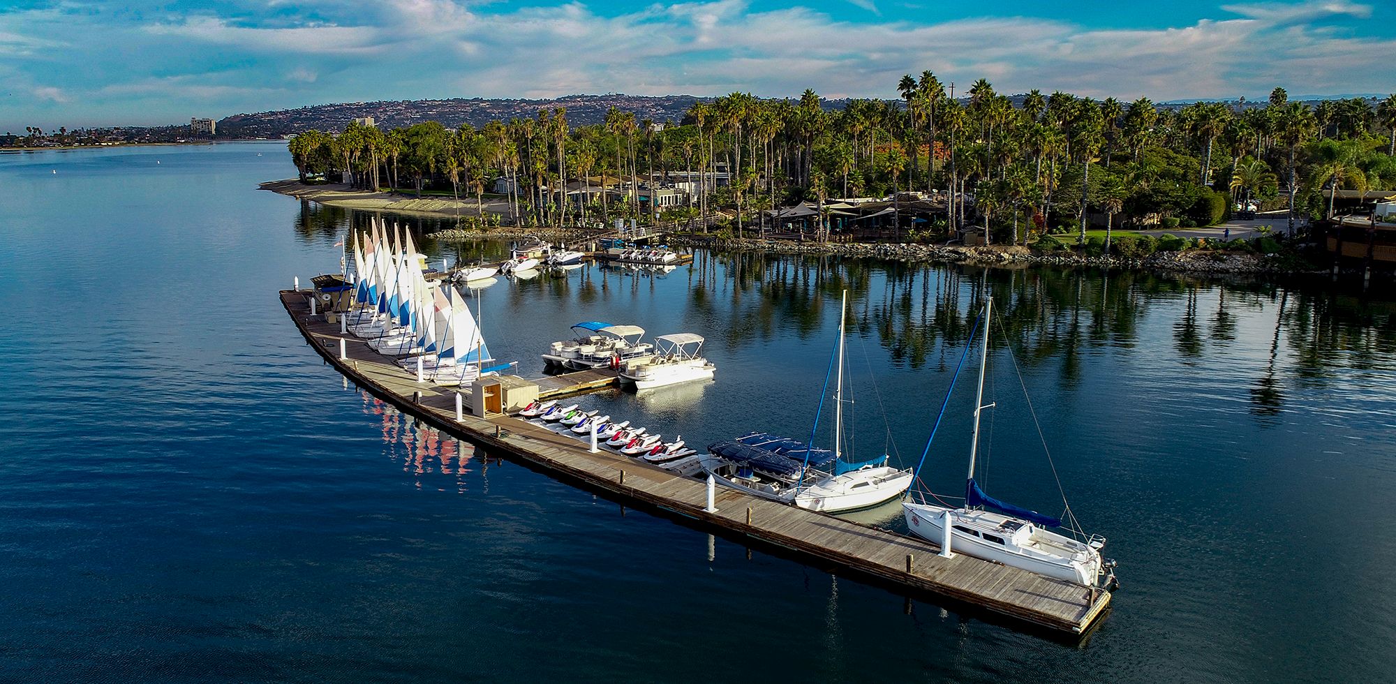 A docked sailboat and yachts sit on a pier beside calm water and a palm-lined waterfront resort under a blue sky.
