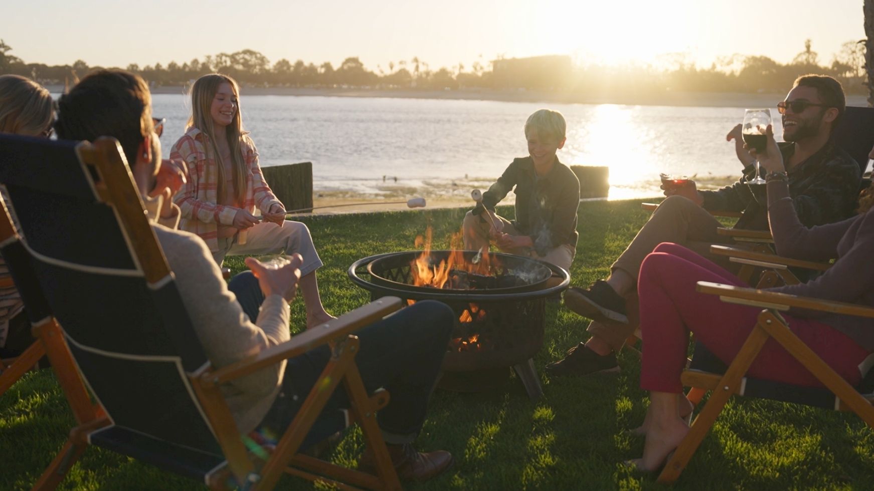 Friends gather around a backyard fire pit by a lakeside at sunset, chatting, toasting drinks, and grilling food together.