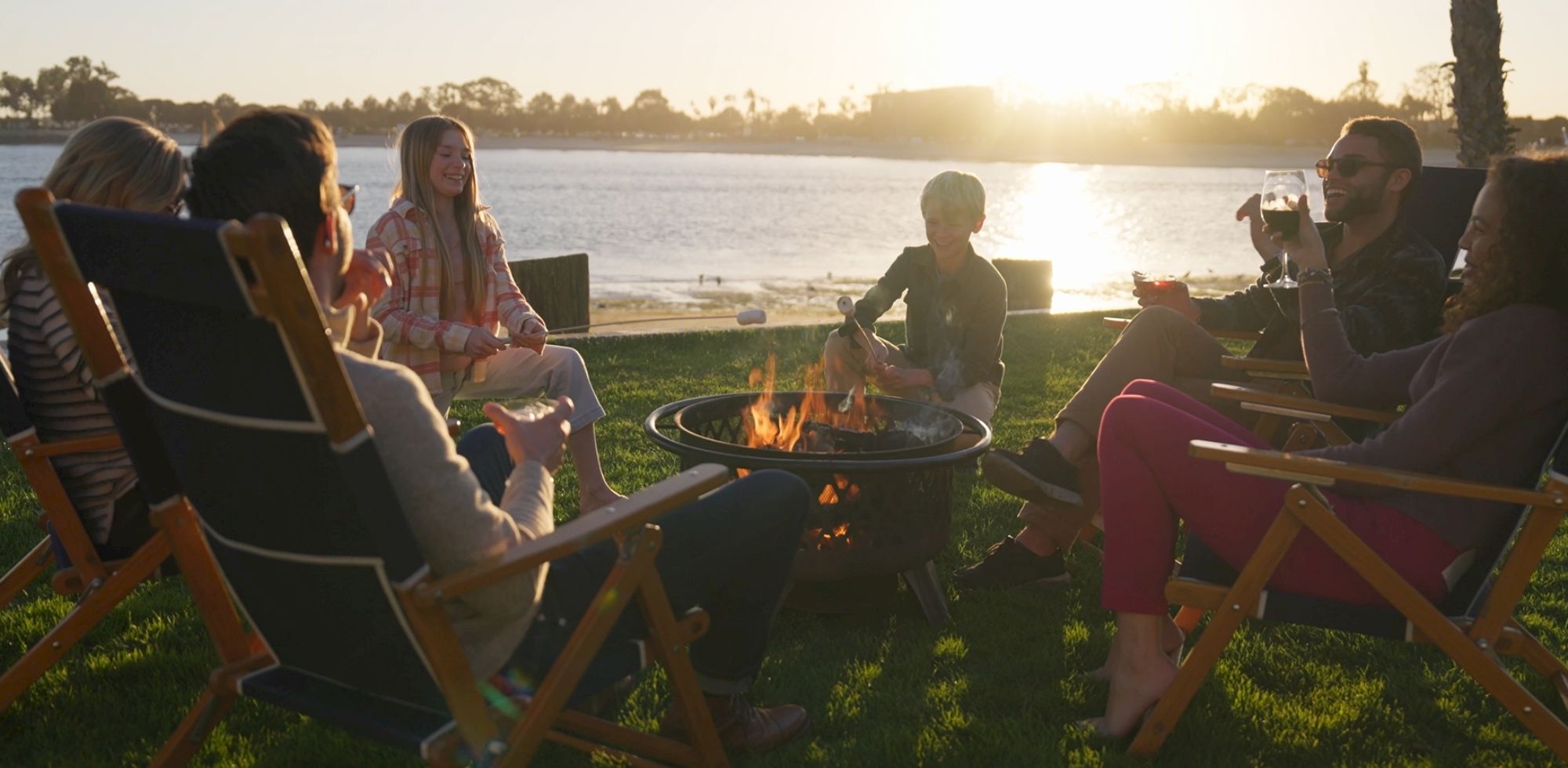 Friends gather around a backyard fire pit by a lakeside at sunset, chatting, toasting drinks, and grilling food together.