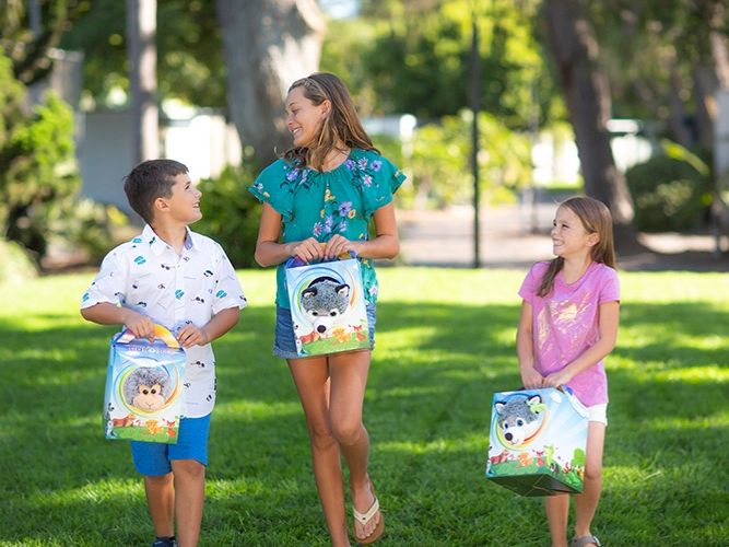 A woman and two kids in a sunny park carry colorful animal lunch bags and smile together.
