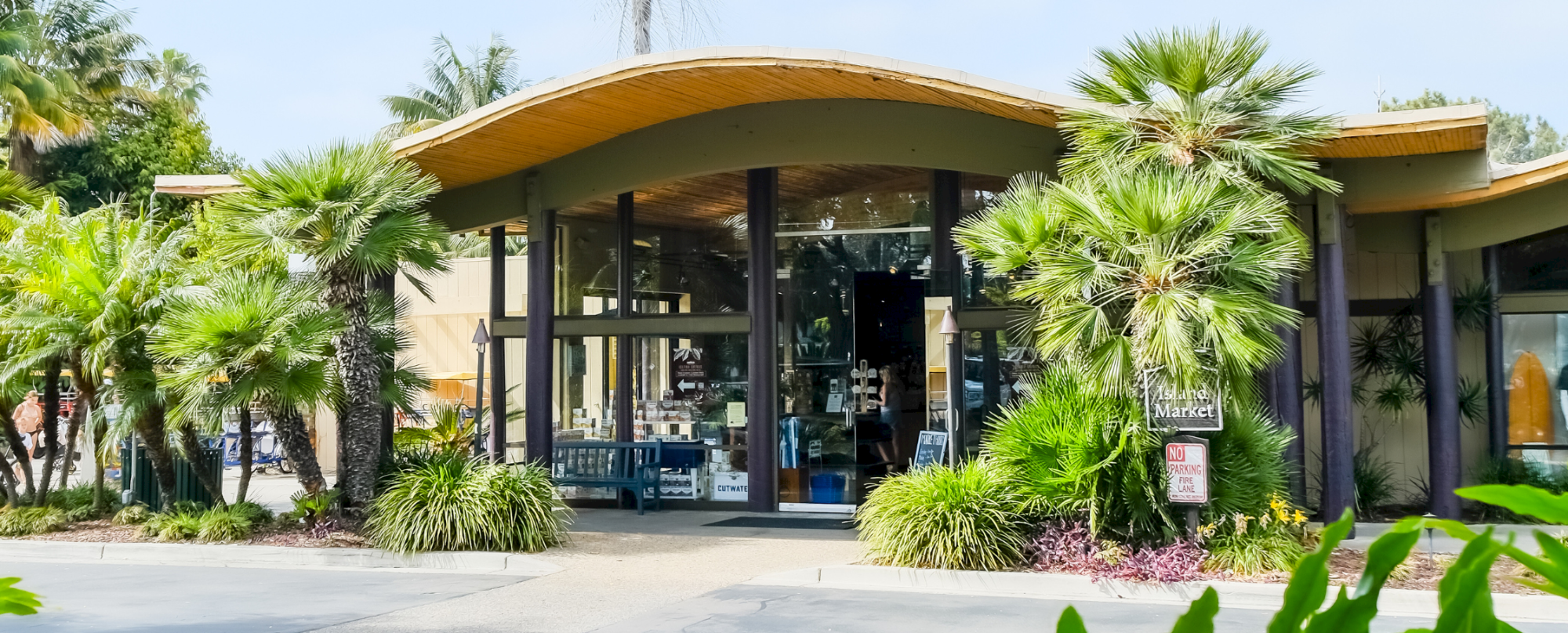 A sunny outdoor entrance to a building with a glass Market sign, framed by palm trees and tropical landscaping.