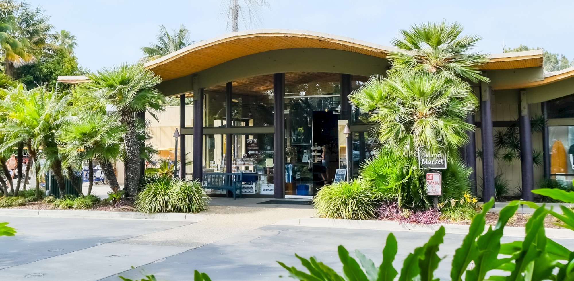 A sunny outdoor entrance to a building with a glass Market sign, framed by palm trees and tropical landscaping.