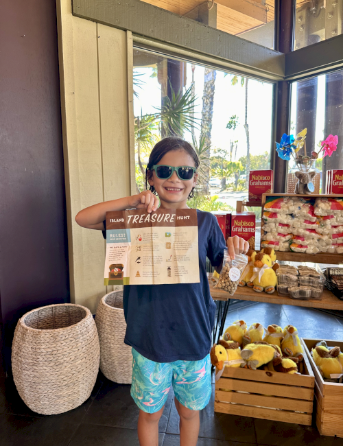 A smiling kid in sunglasses holds an &ldquo;Island Treasure Hunt&rdquo; sheet at a snack-and-souvenir stall by the window.