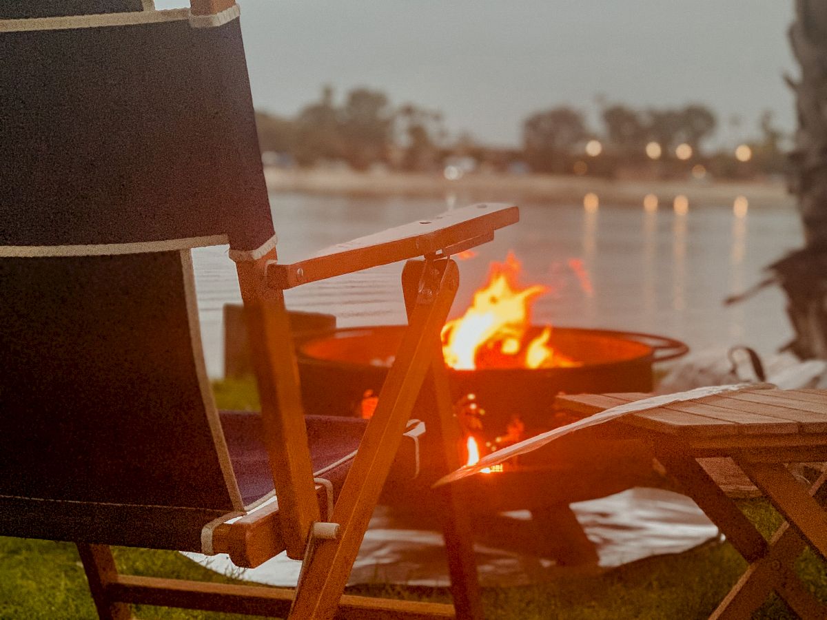 A campfire burns in a metal pit on an outdoor patio by the water as dusk lights the distant skyline.