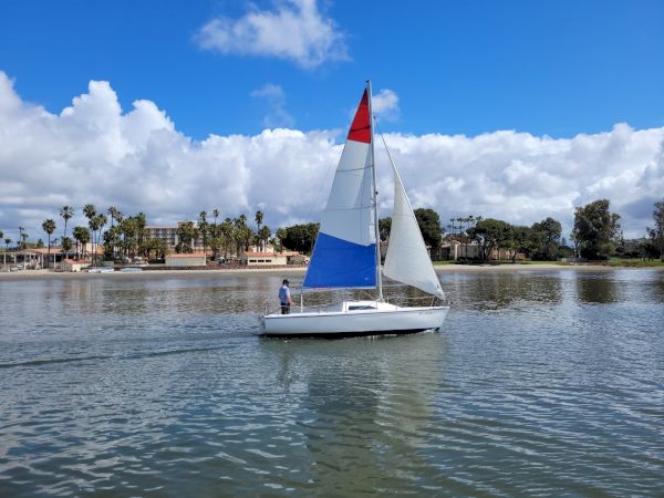 A sailboat with blue and red sails glides on a calm lake beside palm trees under a bright, cloud-filled sky.