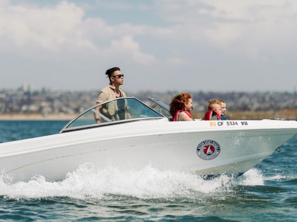A man drives a speedboat while a family rides beside him, kids wearing life vests as the boat speeds across the water.