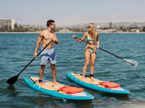 Two people paddleboard on a sunny lake/sea, holding paddles and smiling at each other.