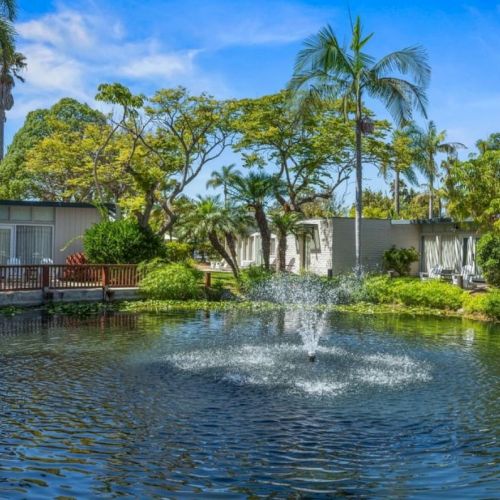 A tropical resort scene: a calm pond with a small fountain in front of palm-lined villas under a blue sky.