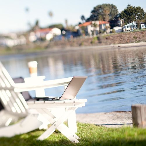 A laptop on a white dockside picnic table by a calm river, with houses and trees in the sunny background.