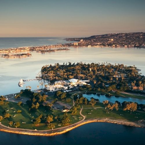 Aerial view of a palm-covered island resort in a calm bay at sunset, with docks, lawns, and a coastline city nearby.