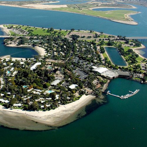 Aerial view of a small resort island with sandy beaches, docks, boats, and a bridge over a wide river.
