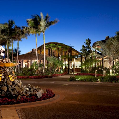 A luxury resort at dusk with palm trees, lit buildings, and a stone fountain in a landscaped garden.
