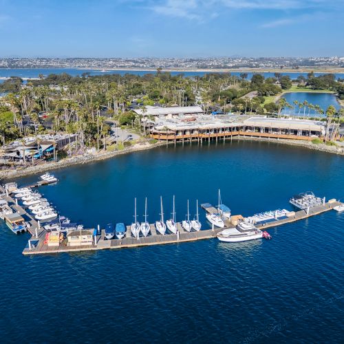 Aerial view of a marina with yachts moored at docks, palm-lined waterfront, and a long pier over blue ocean.