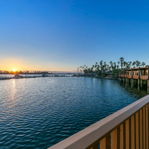 Golden sunset over a calm marina, with docked boats, palm-lined shore, and a wooden walkway by the water.