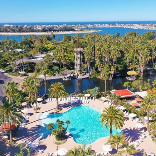 An aerial view of a tropical resort with a large turquoise pool, palm trees, and a coastline in the distance.