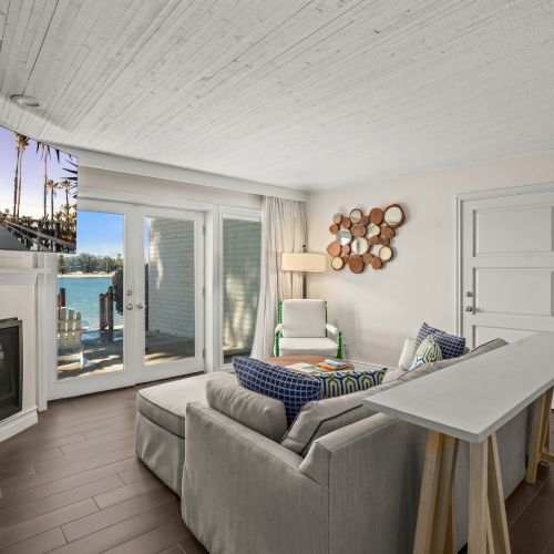 Bright living room with gray sofa, lit fireplace, round wall art, and big windows showing palm trees by the water.