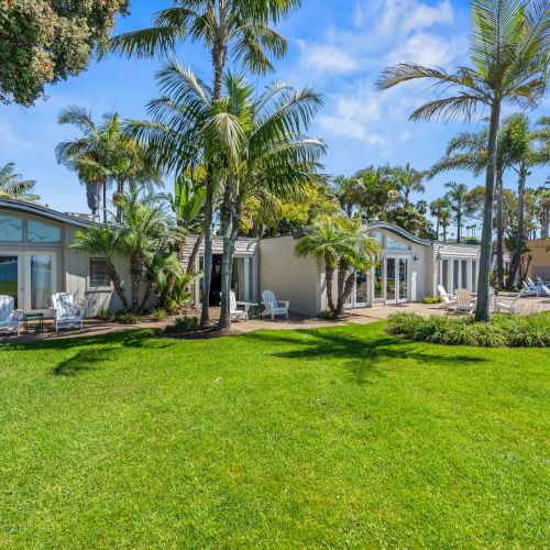 Tropical resort courtyard with palm trees, manicured lawn, and white bungalow rooms with patio chairs under a bright blue sky.