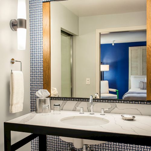 Modern bathroom with a marble vanity, double faucets, large mirror, towel bar, and blue patterned walls showing a bedroom beyond.