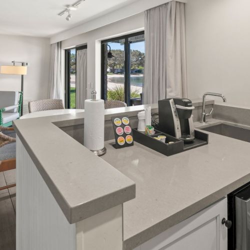 Modern kitchen with gray countertops, sink, coffee maker, and chairs by large windows with a garden view.