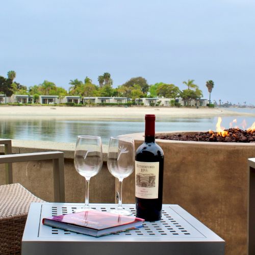 A lakeside patio table set with wine bottle and glasses beside a roaring fire pit, facing a calm beach and palm-lined shore.