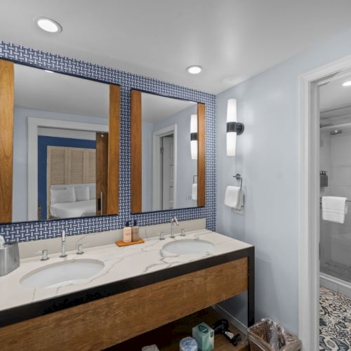 Modern bathroom with dual sinks, blue patterned wall, large mirror, and a glass shower with patterned tile flooring.