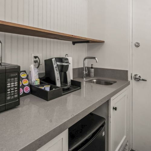 A compact hotel kitchenette with a microwave, coffee machine, stocked water bottles, and a stainless sink and soap dispenser.