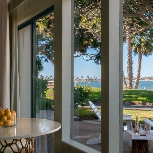 A sunny oceanfront patio: a round table with lemons by open doors, white chairs, and palm trees overlooking the blue sea.