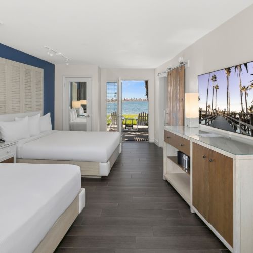 A bright hotel room with two white beds, ocean view through windows, and a TV with palm trees on the wall.