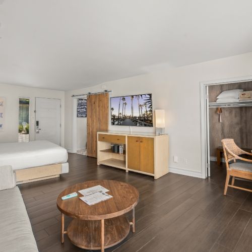 A modern hotel room with a bed, sofa, round coffee table, small kitchenette, and beach-themed wall art.