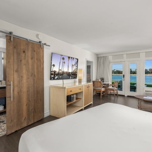 Modern hotel room with a bed, vanity and wooden door, opening to a balcony view of a calm lake.