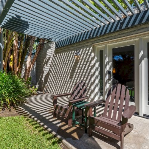 Outdoor patio with striped pergola, brick wall, and wooden rocking chairs on a sunlit walkway.