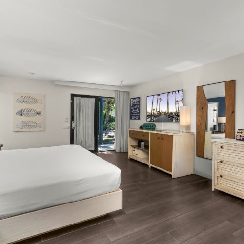 Bright hotel room with a white bed, dresser, coffee maker, wall art, and a sliding door to a poolside view.