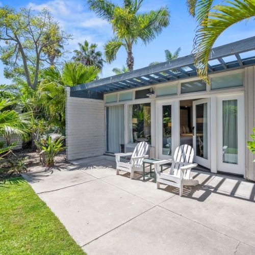 A sunny tropical patio beside a modern home, with palm trees, lush greenery, and white outdoor chairs by the sliding glass doors.