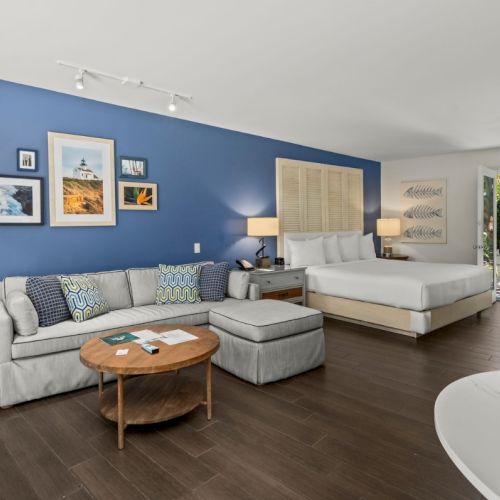 A bright coastal-style bedroom with a blue accent wall, framed photos, a sofa, a round coffee table, and a neatly made bed, inside.