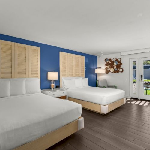 A modern bedroom with two white beds, blue accent wall, shutters, and an open door to a patio through large windows.